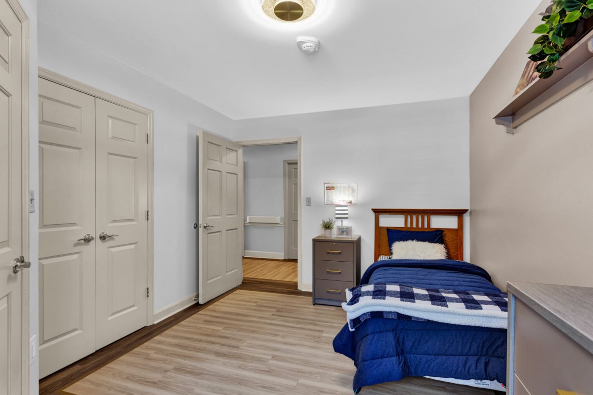 A cozy bedroom with a single bed featuring blue bedding, a small dresser with a lamp, and wooden floors.
