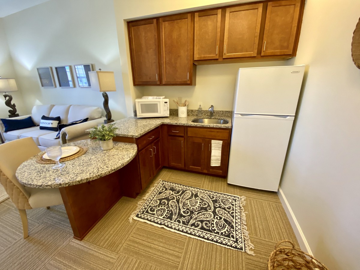 A cozy apartment kitchen with wooden cabinets, a refrigerator, microwave, and a patterned rug on the floor.
