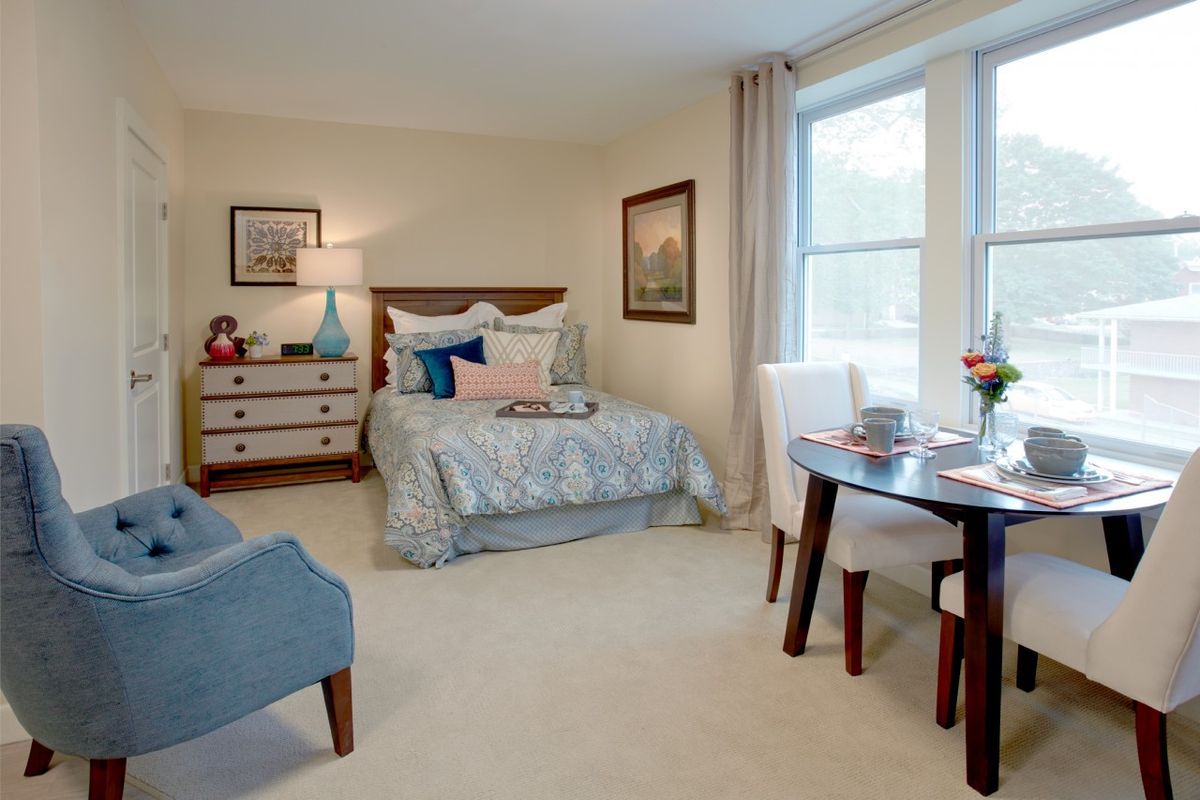 A cozy and well-decorated bedroom featuring a comfortable bed, a dresser with a lamp, and a small dining table by the window.