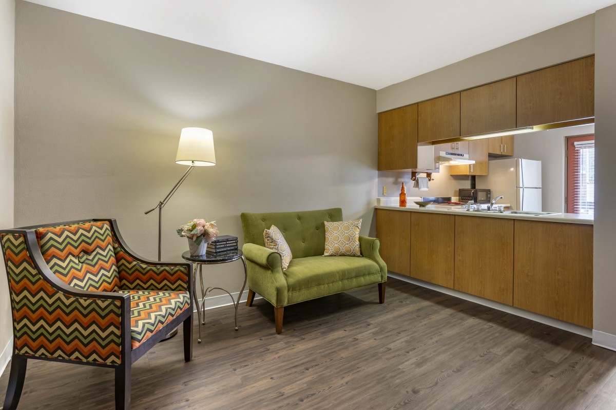 A cozy living space with a green sofa, patterned armchair, and a kitchenette in the background.