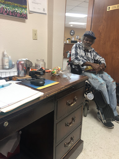 A man seated in a wheelchair in an office setting, accompanied by a desk and various items.