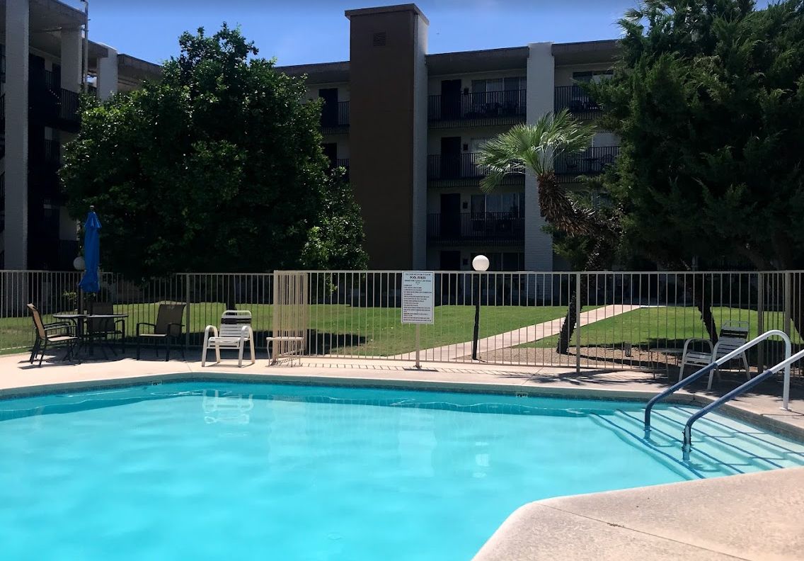A clear day at an apartment complex pool surrounded by trees and a fence.