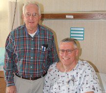 Two men in a hospital room, one in patient attire and the other standing beside him.