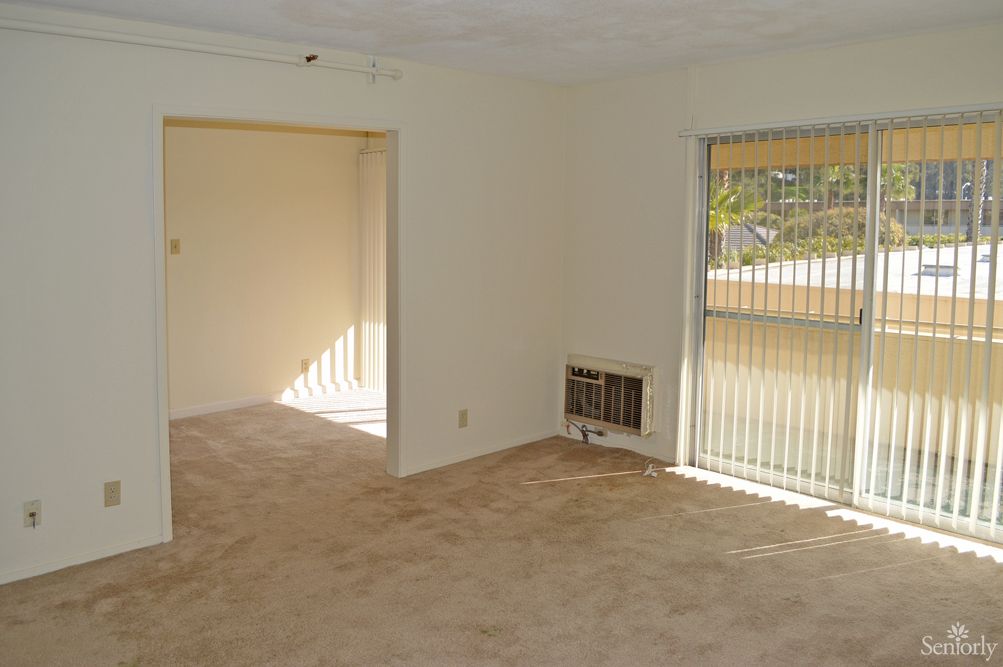 An empty room with carpet flooring, windows with vertical blinds, and a doorway leading to another area.