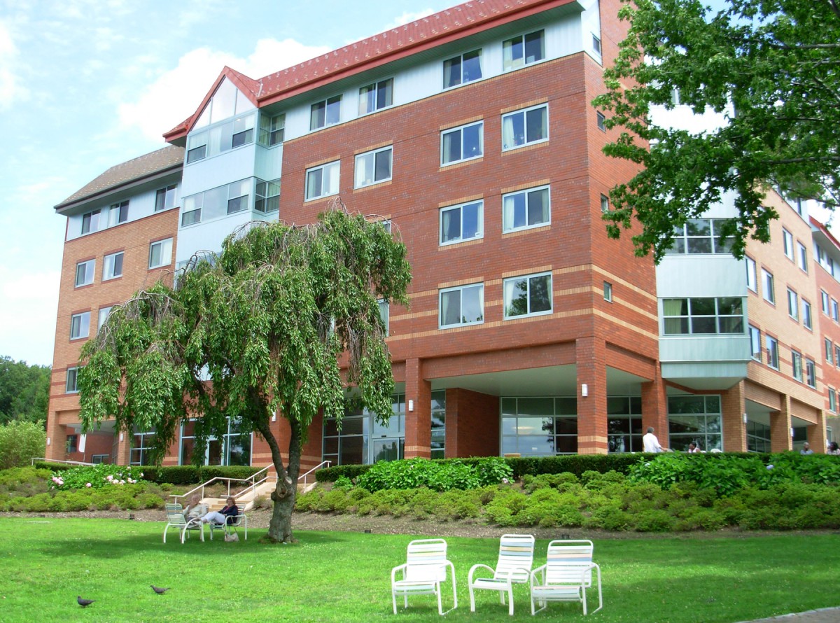A multi-story brick residence building with chairs on the lawn and trees surrounding it.