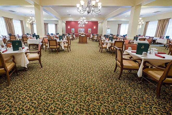 A spacious dining area with neatly arranged tables and chairs, featuring elegant chandeliers and a patterned carpet.
