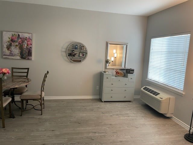 A cozy living room with a dresser, decorative items, and a dining set next to a large window with blinds.
