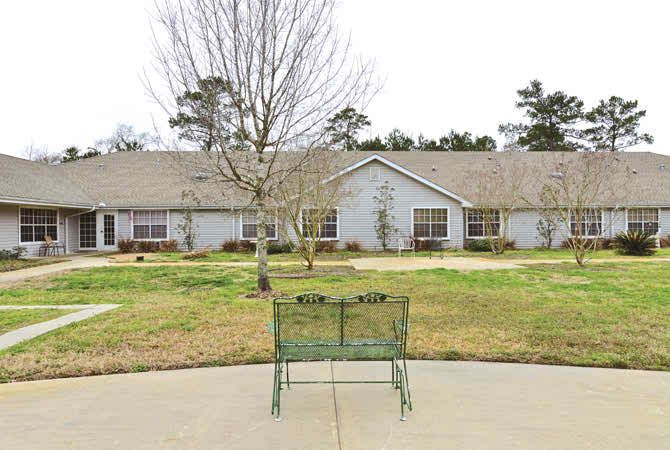 A view of a single-story building with a bench in the foreground and trees in the yard.