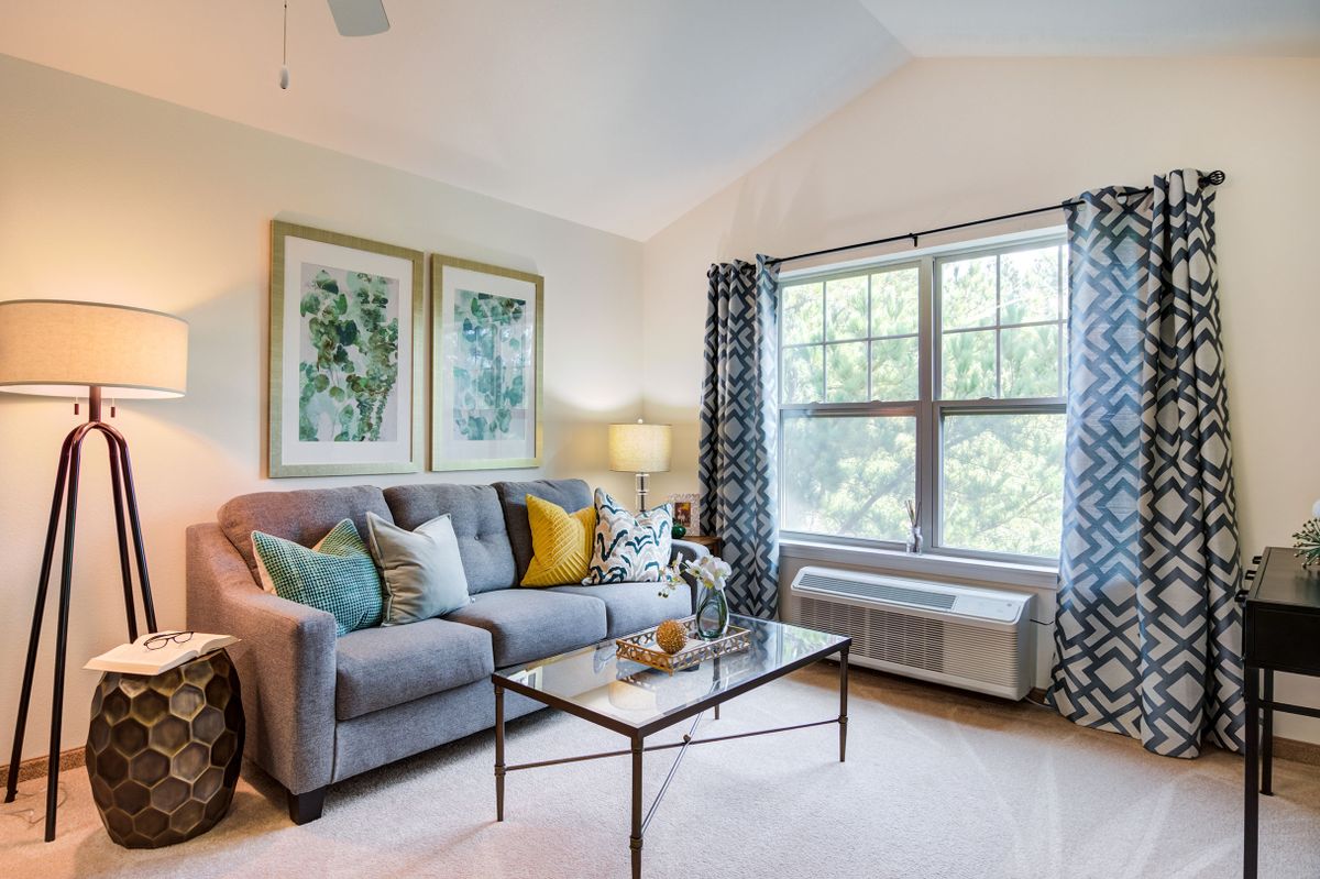 A cozy living room featuring a gray couch adorned with colorful pillows, a glass coffee table, and decorative artwork on the wall. Light flows through the window with patterned curtains.