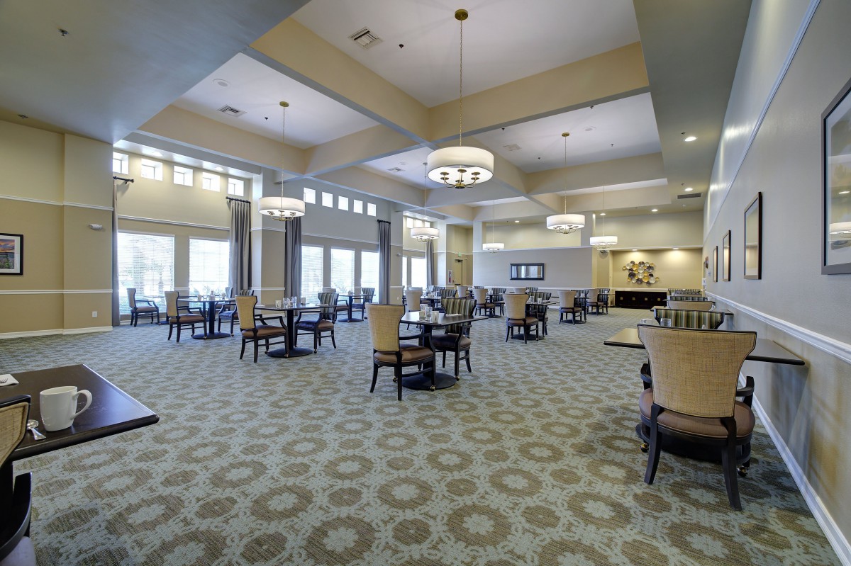 A spacious dining room with elegant chandeliers, tables, and chairs arranged on a patterned carpet.