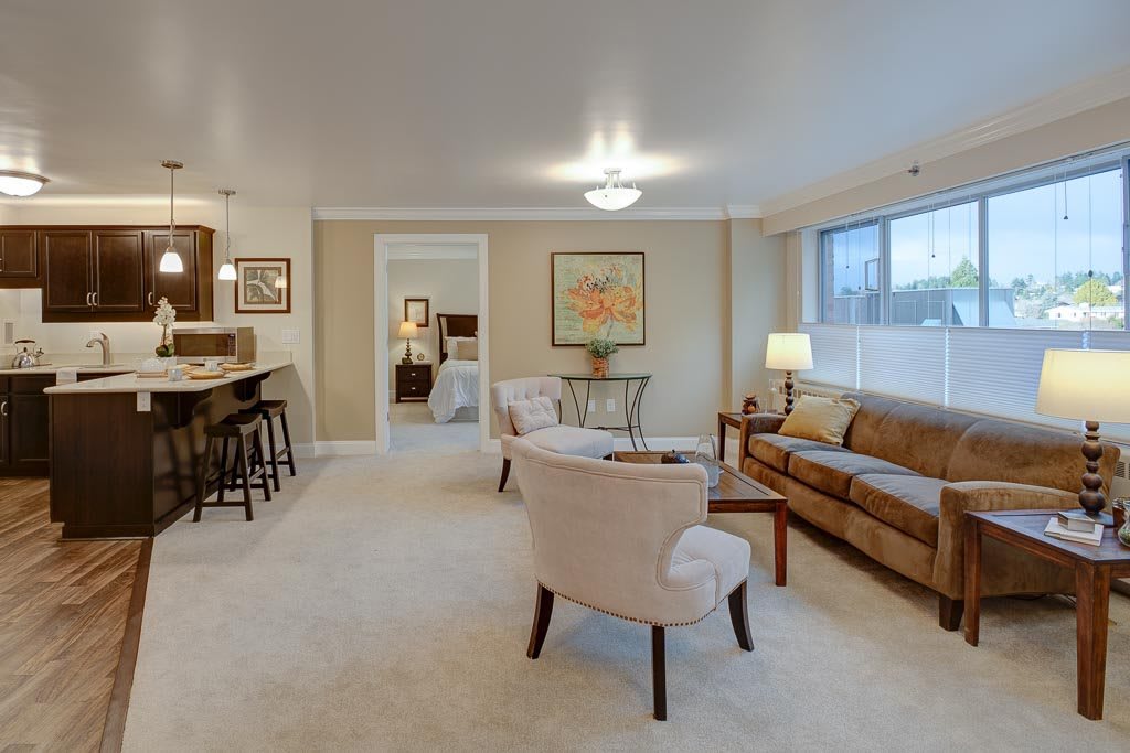 A cozy living room with neutral tones, featuring a sofa and chairs around a coffee table, complemented by a large window for natural light.