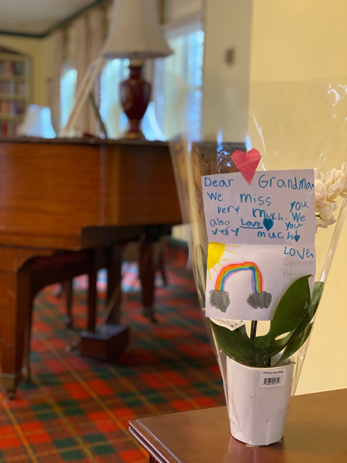 A flower bouquet with a handwritten note stands in front of a grand piano in a cozy room.