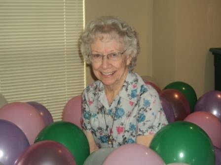 An elderly woman happily surrounded by colorful balloons indoors.
