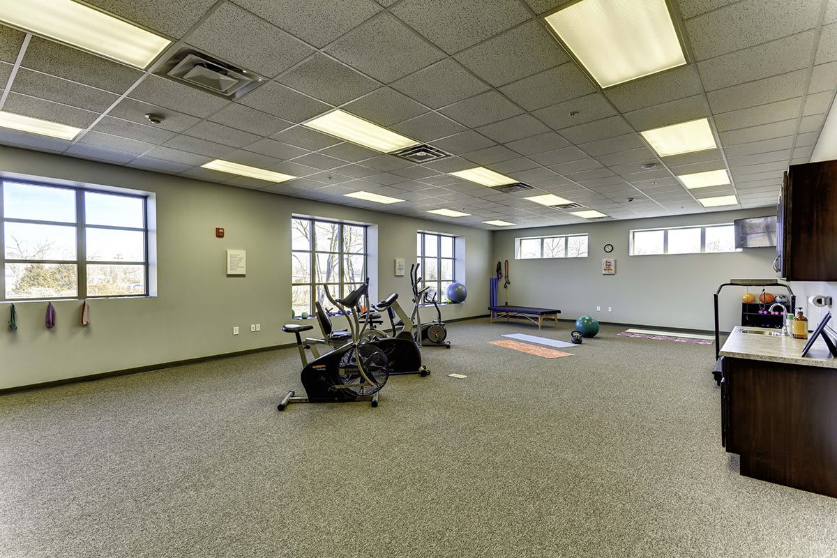 An empty gym room featuring stationary bikes and yoga mats, with large windows providing natural light.