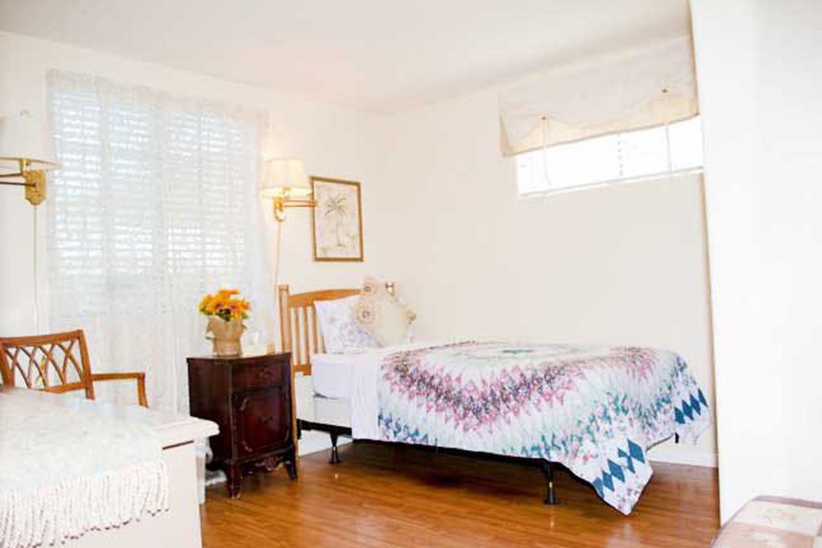 A cozy bedroom with a single bed adorned with a colorful quilt, surrounded by warm lighting and wooden floors.