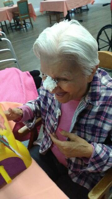 An elderly woman smiling with cake on her nose during a celebration.
