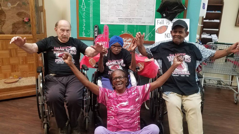 A joyful group of people posing together, some in wheelchairs, all smiling and wearing themed t-shirts indoors.