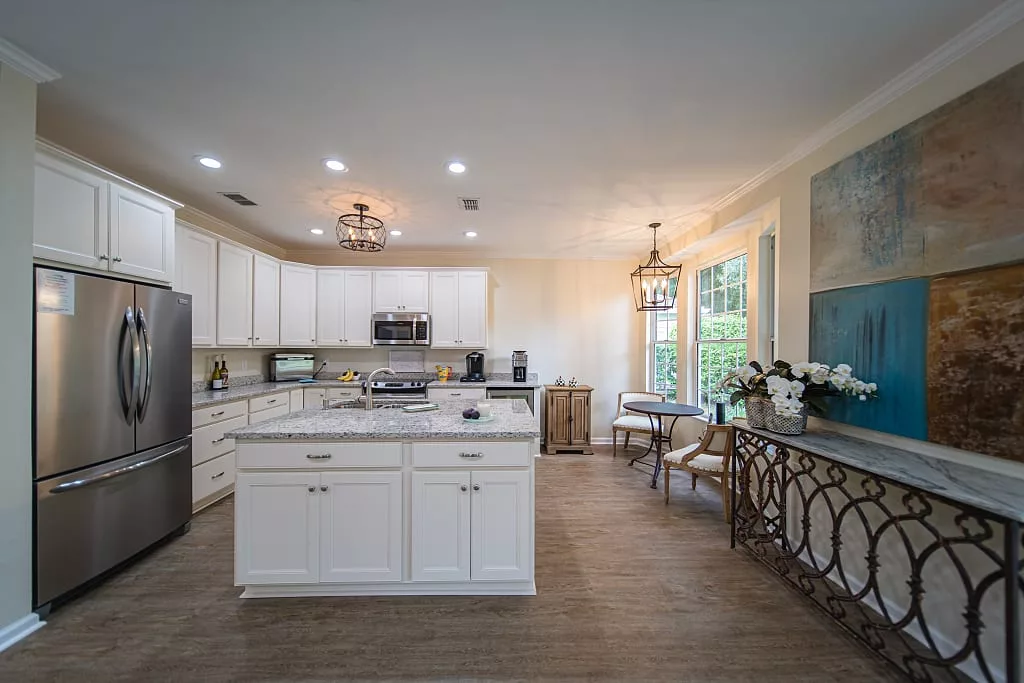 A modern kitchen featuring white cabinets, an island, wood flooring, and a connected dining area with stylish lighting fixtures.