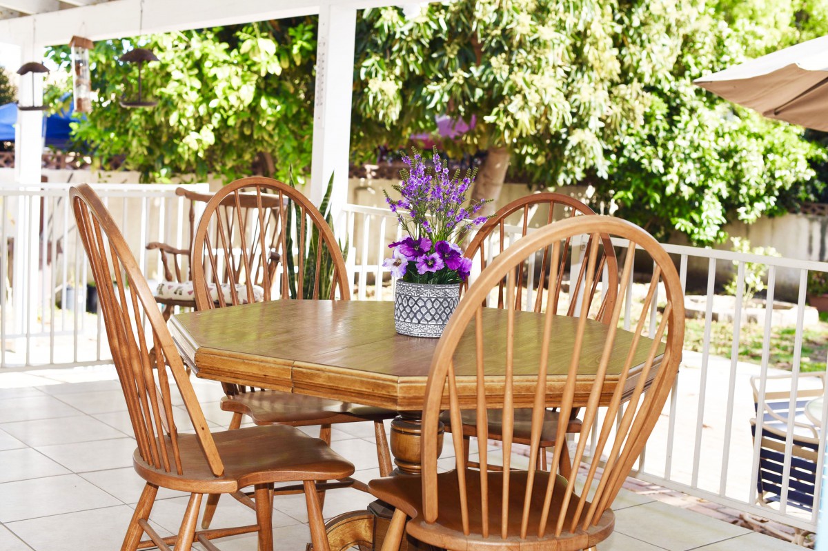 A cozy outdoor patio with a wooden table, surrounded by chairs and adorned with a vase of flowers in sunlight.