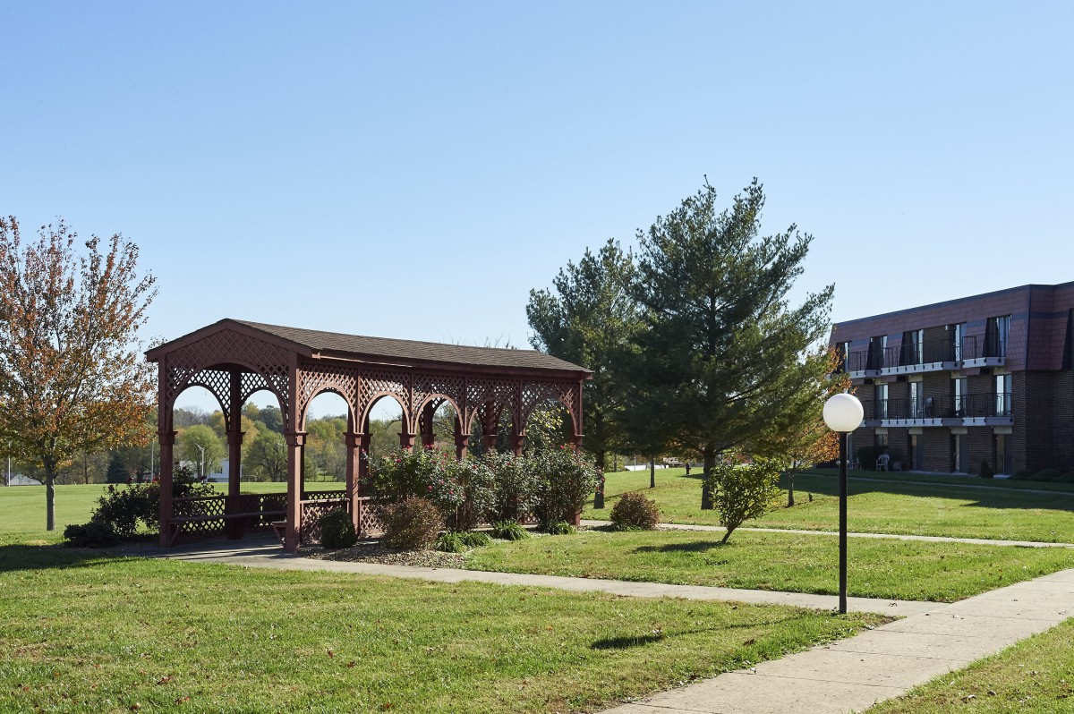 A scenic view of a park featuring a wooden gazebo, surrounding trees, and a well-maintained grassy area under a clear blue sky.