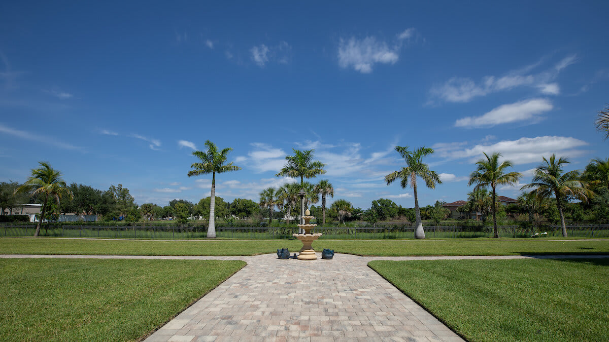 A tranquil landscape featuring a paved pathway leading to a fountain, surrounded by palm trees and a lush lawn under a clear blue sky.