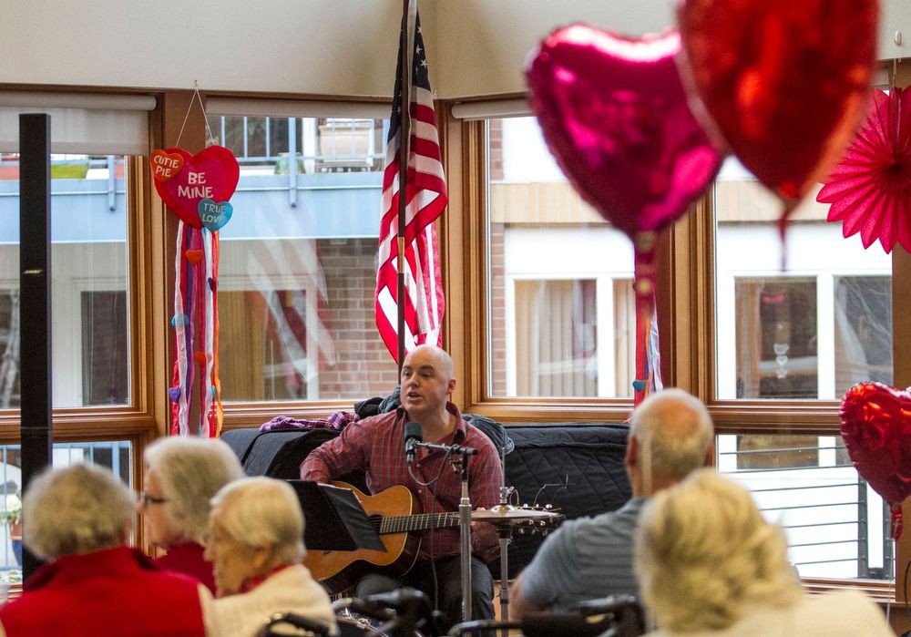 A musician performs with a guitar for an audience in a decorated room with heart-shaped balloons and an American flag.