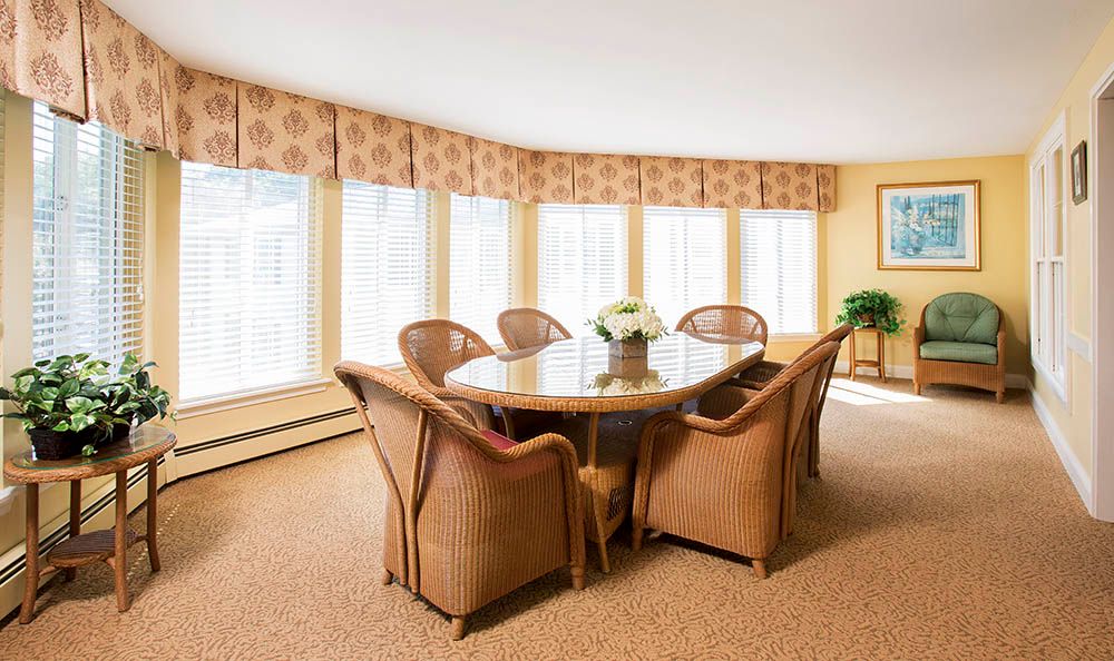 A well-lit dining room with a wooden table surrounded by six chairs, featuring large windows and patterned curtains.