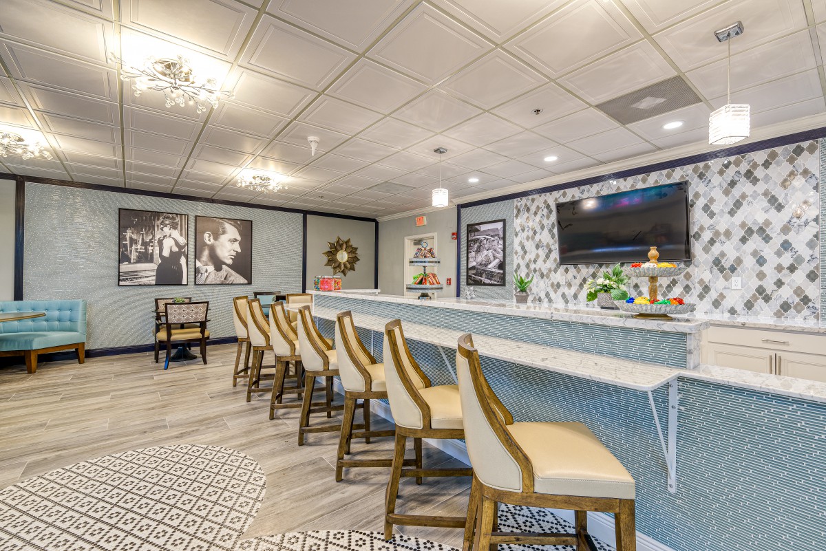 A stylish bar interior featuring a long counter with upholstered chairs, decorative lighting, and wall art.