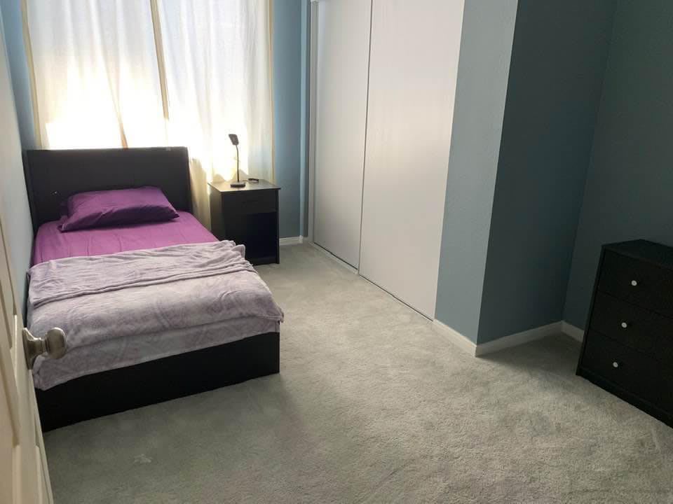 A tidy bedroom featuring a bed with purple bedding, a nightstand with a lamp, and a closet against the wall.