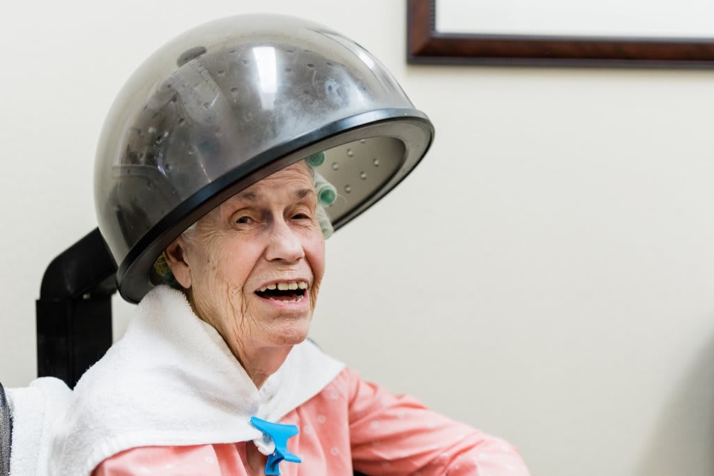 An elderly woman smiling while sitting under a hair dryer at a salon.