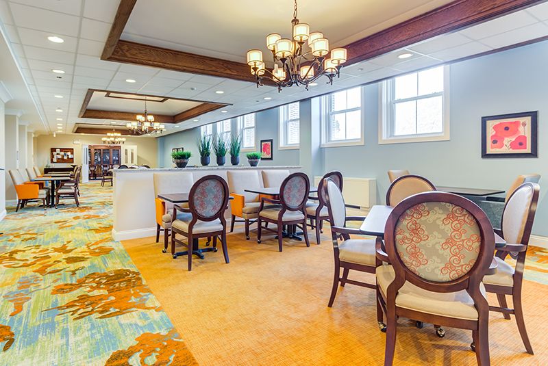 A brightly lit dining area featuring elegant chandeliers, comfortable chairs, and tables on a carpeted floor.