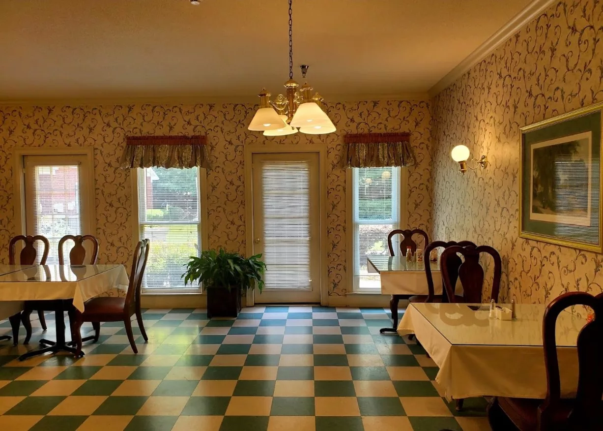 A vintage-style dining room with checkered flooring, decorated with wallpaper and featuring tables and chairs under warm lighting.