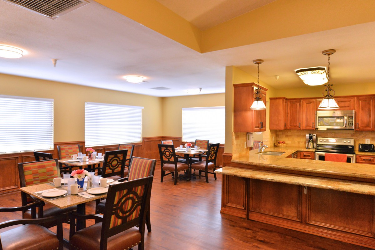 A cozy dining room with neatly arranged tables and chairs, adjacent to a kitchen area with wooden cabinets.