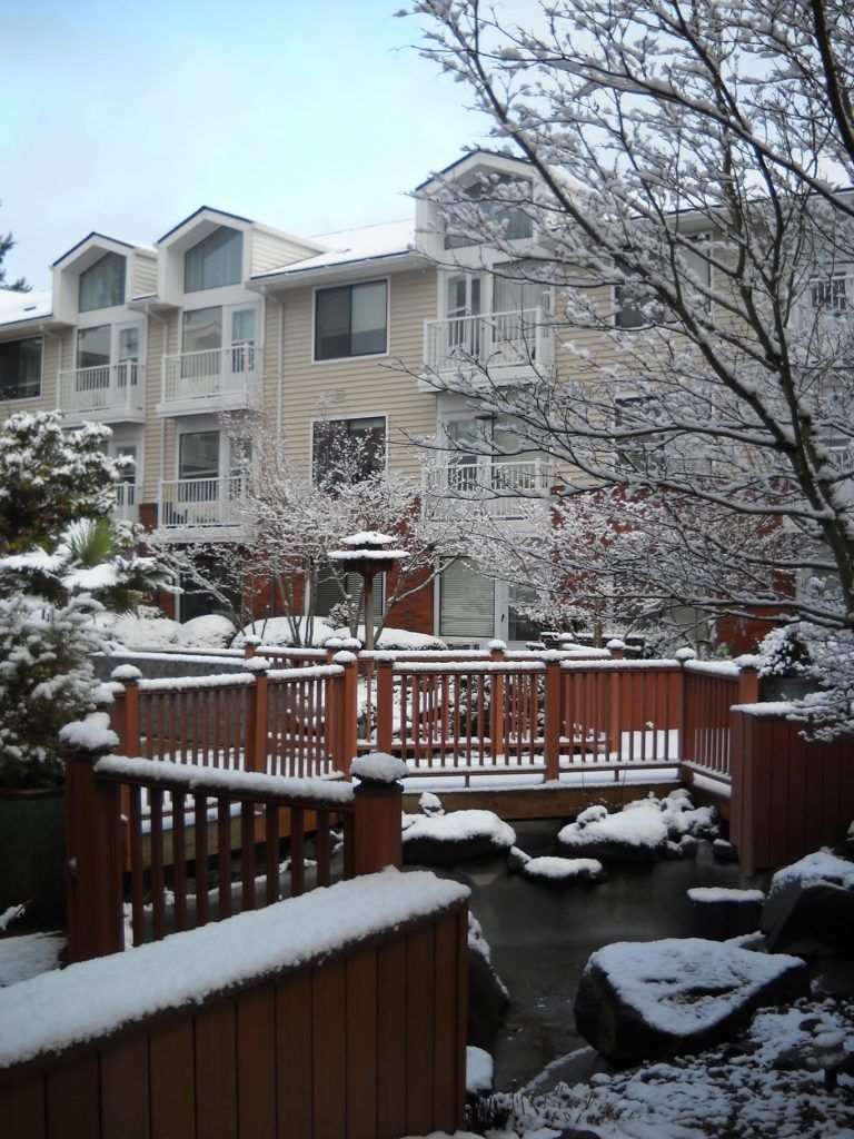 A snowy winter scene featuring an apartment building with a wooden deck and snow-covered trees.
