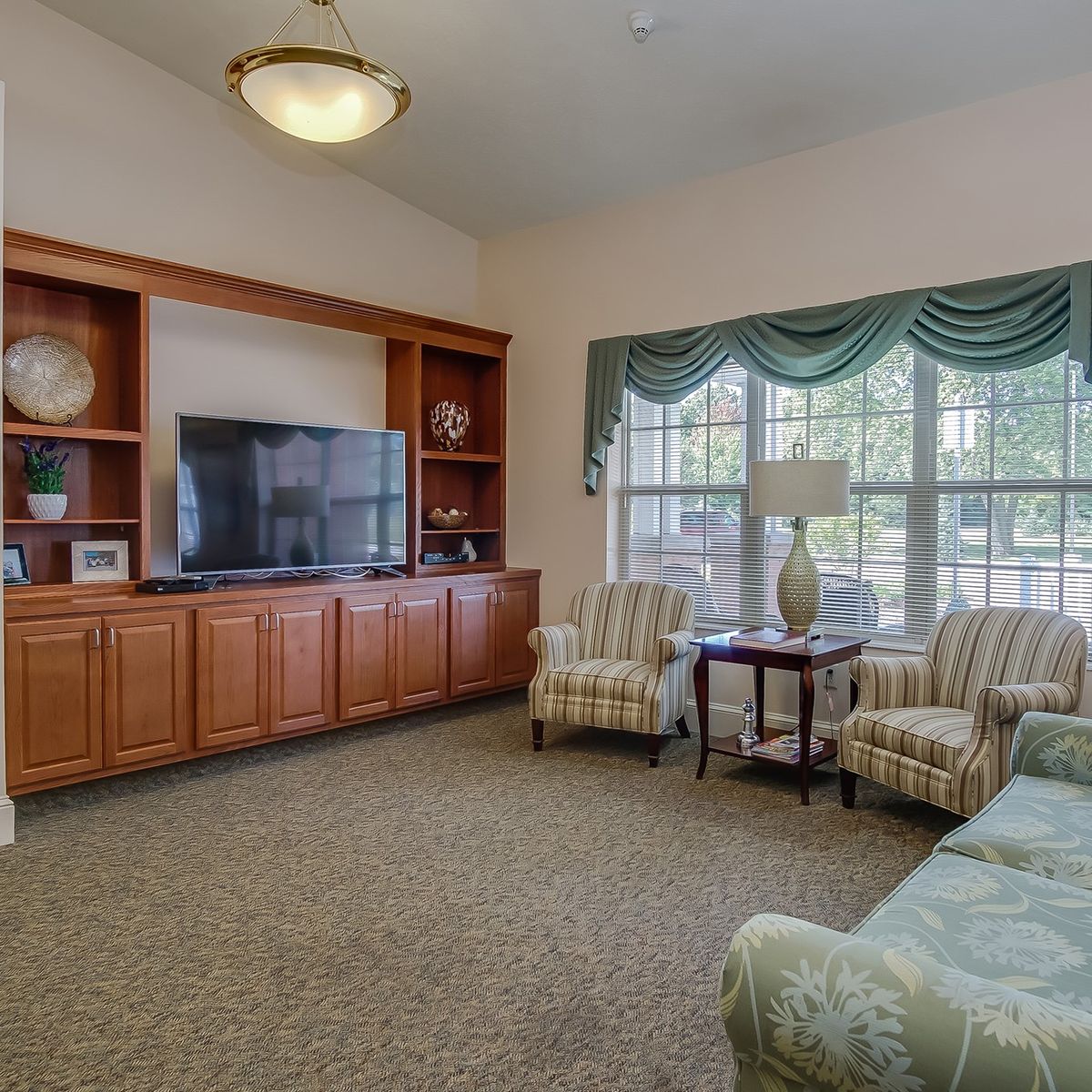 A cozy living room with a large television, sofa, and armchairs arranged around a coffee table, featuring large windows and elegant curtains.