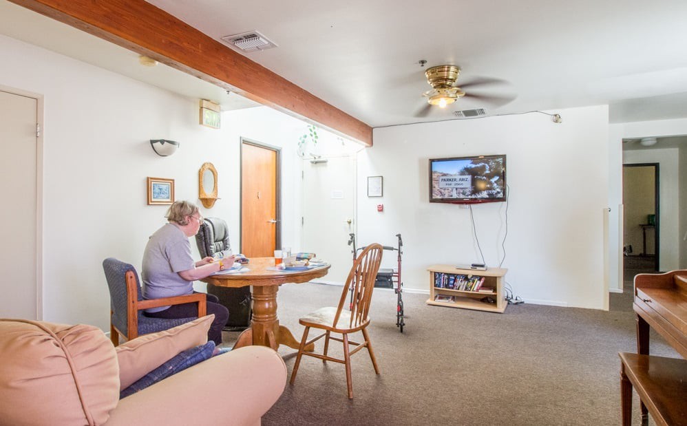 A person seated at a table in a cozy living room with a TV and ceiling fan.