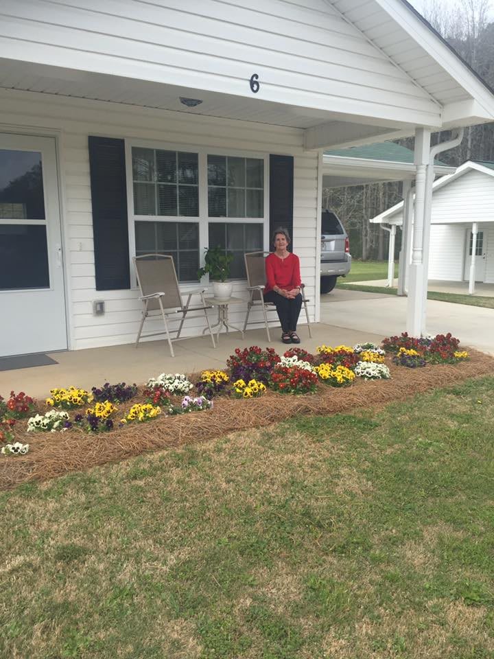 A person sitting on the porch of a house, surrounded by a garden with vibrant flowers.