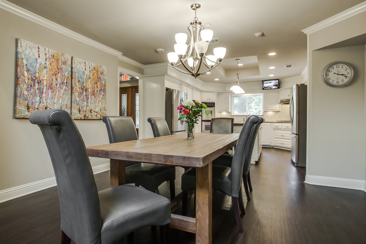 A modern dining room with a wooden table, elegant chairs, and a chandelier, opening into the kitchen area.