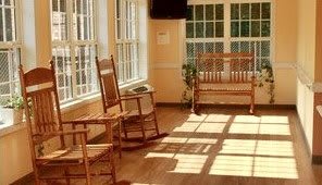 A cozy sunroom with wooden rocking chairs and a bench, illuminated by sunlight streaming through the windows.