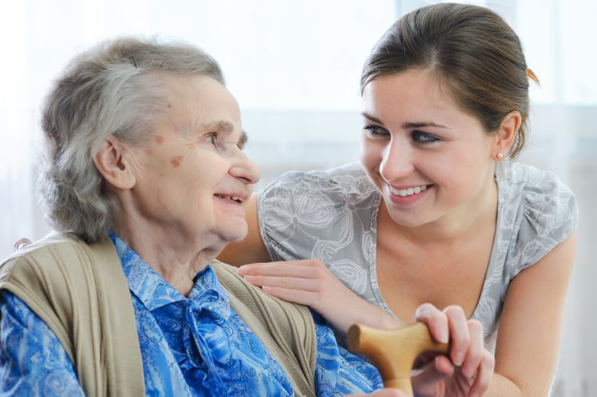 A young woman smiling and supporting an elderly woman in a caring interaction.