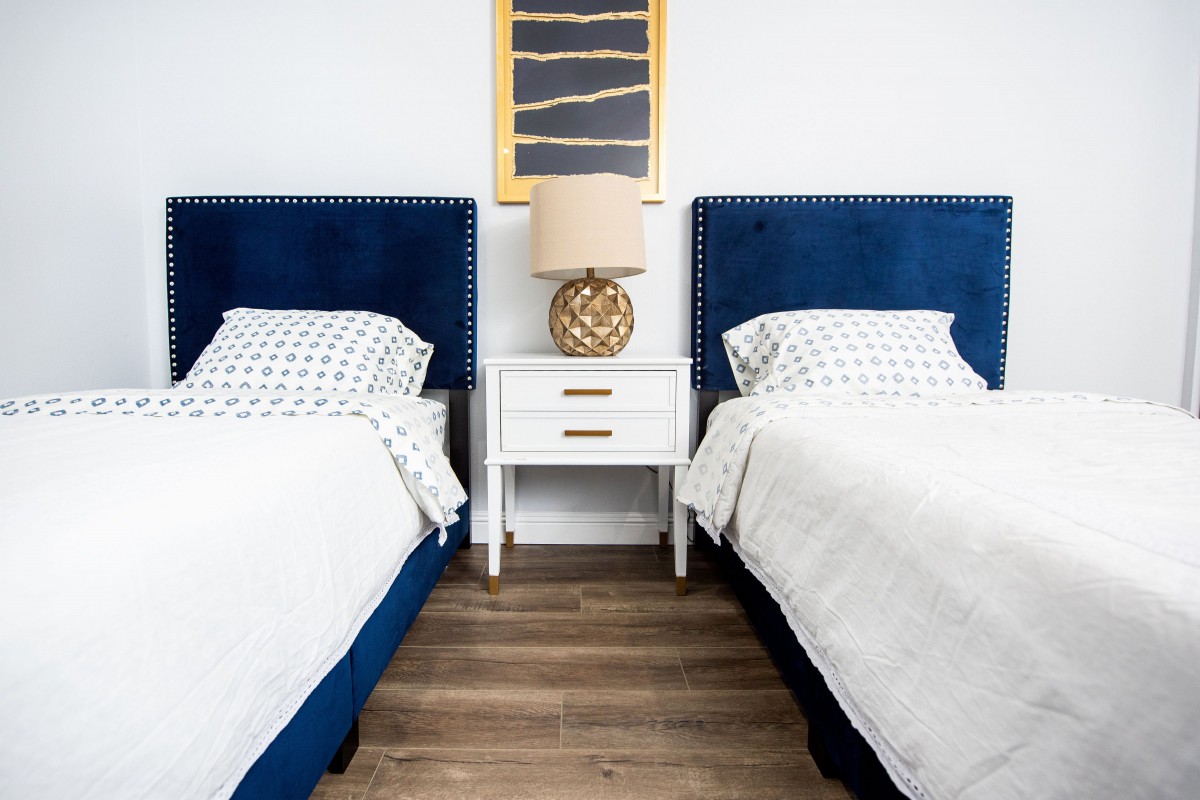 A modern bedroom featuring twin beds with blue headboards, a central nightstand, and framed art above.