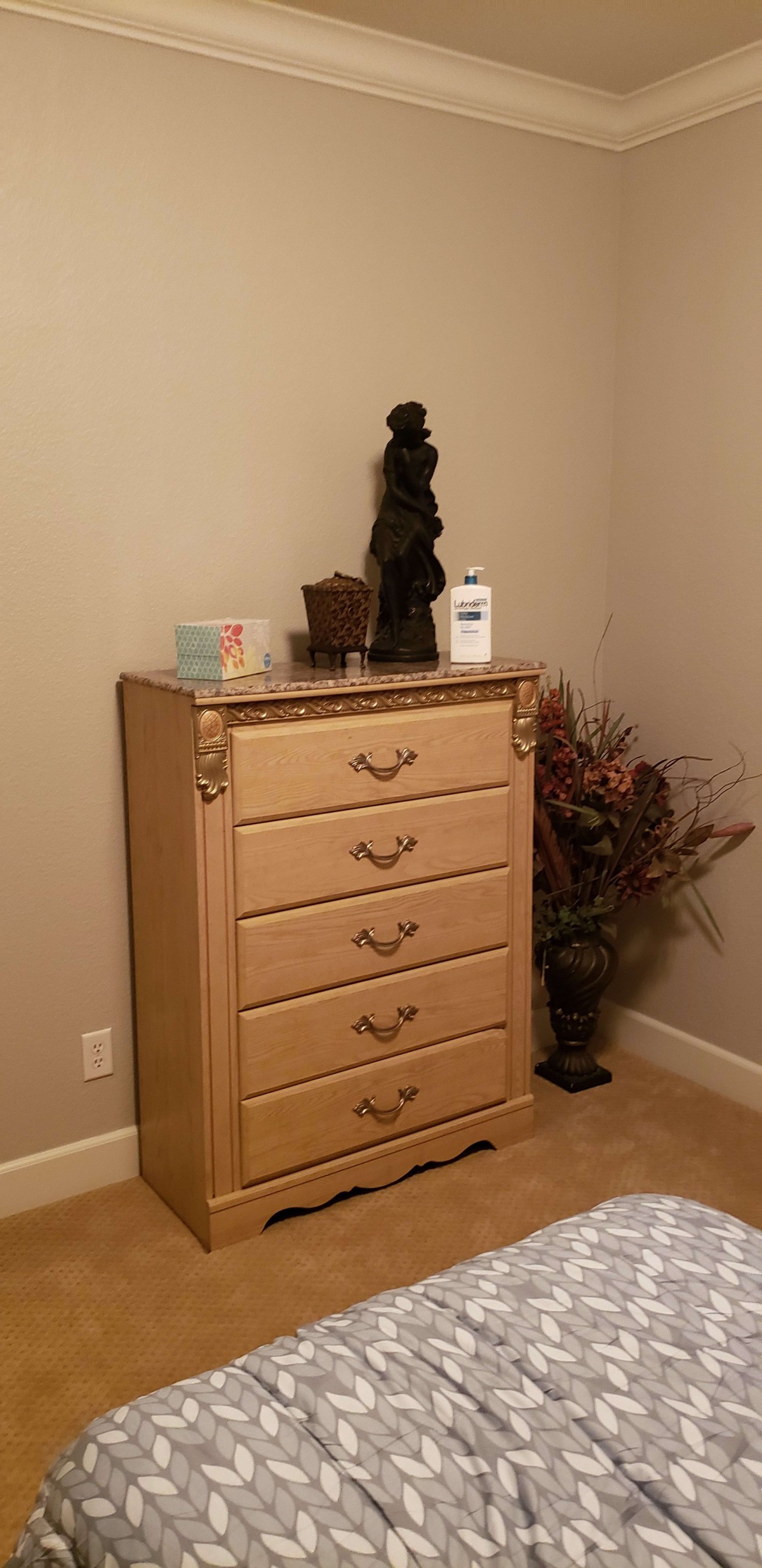 A wooden dresser with five drawers, adorned with a decorative statue and vase, situated in a corner of a room.