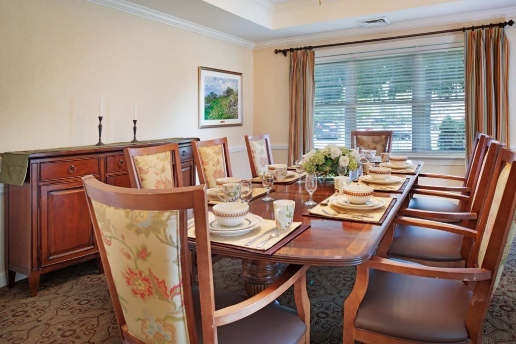 A beautifully set dining room table with elegant place settings and upholstered chairs, situated near large windows with draped curtains.