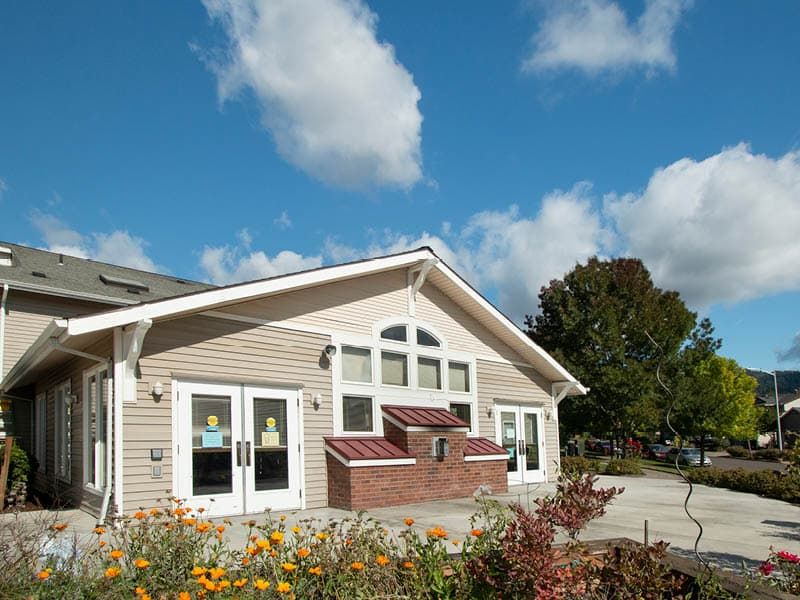 A modern house with large windows under a partly cloudy sky, surrounded by trees and flowers.