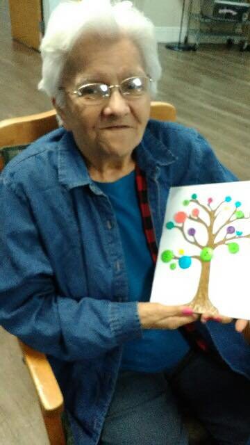 An elderly woman holding a colorful painting of a tree.