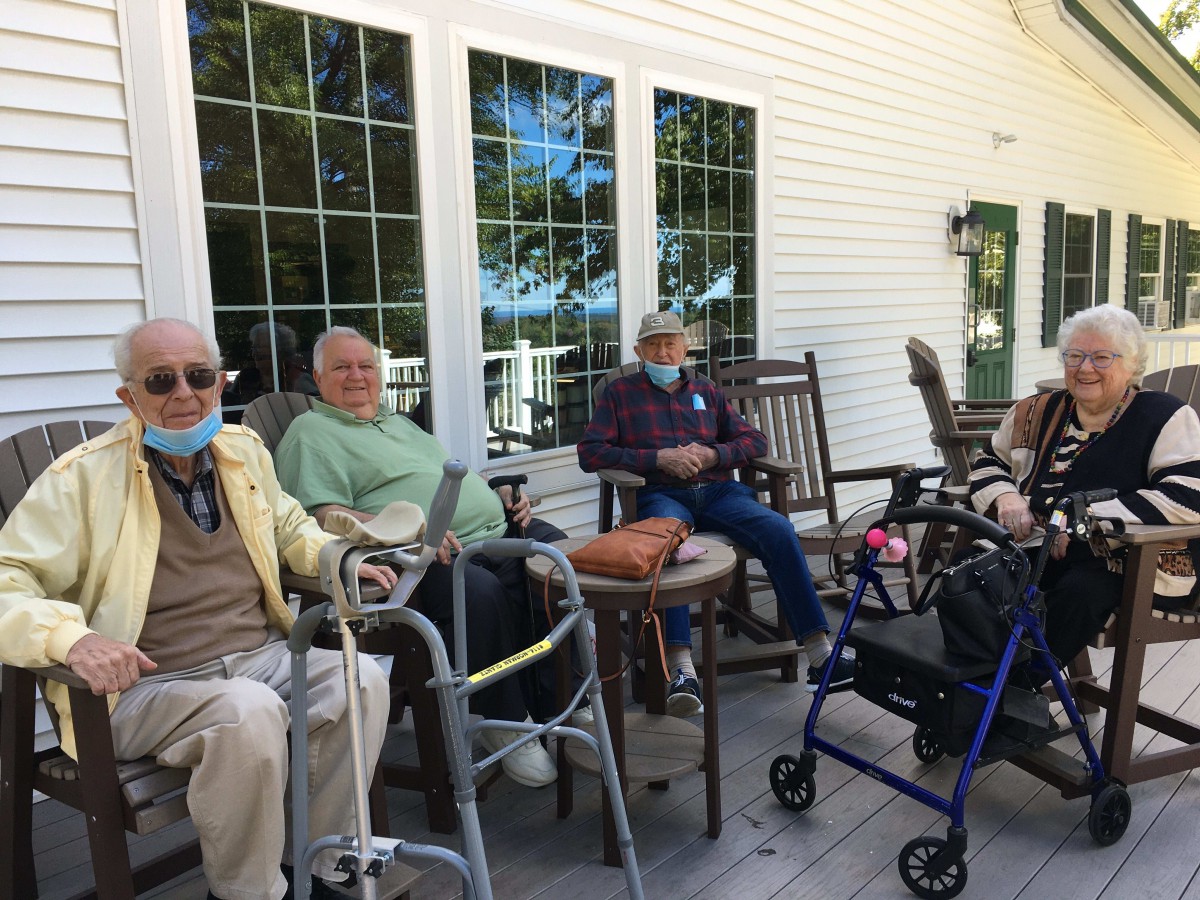 A group of elderly individuals sitting on a porch, some using walkers and wearing masks.