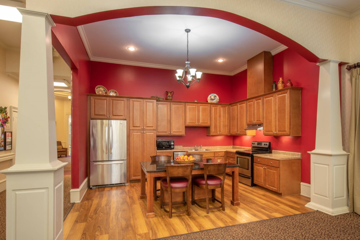 A cozy kitchen with wooden cabinets and red walls, featuring modern appliances and a dining table in the center.