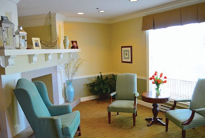 A cozy living room with two chairs, a small table with flowers, and a decorative fireplace.