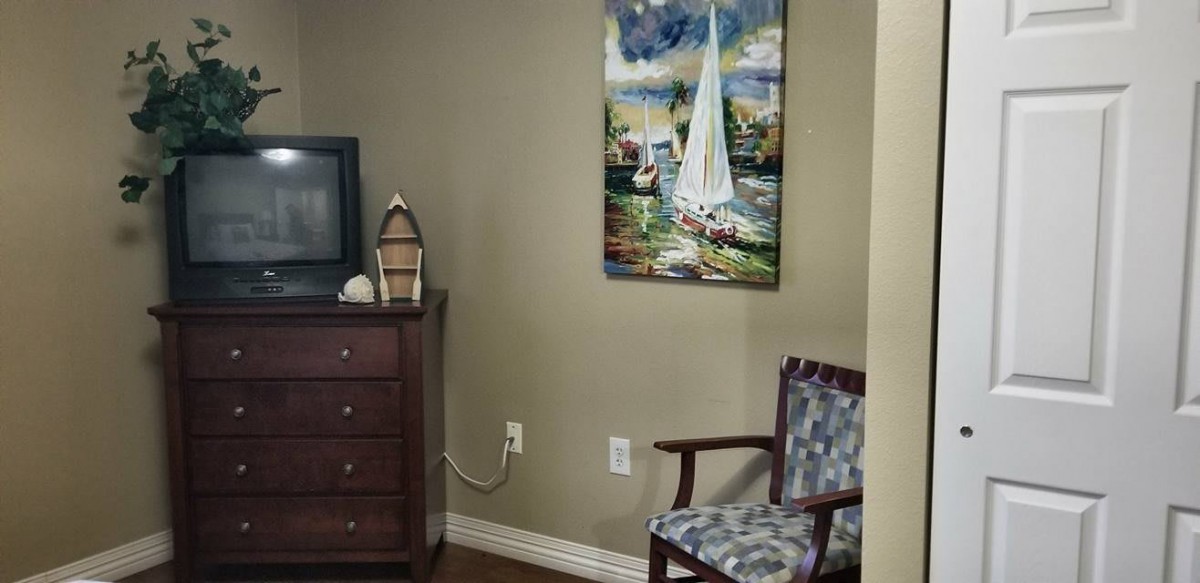 A cozy corner with a television atop a dresser, a decorative lamp, and a sailboat painting above an upholstered chair.