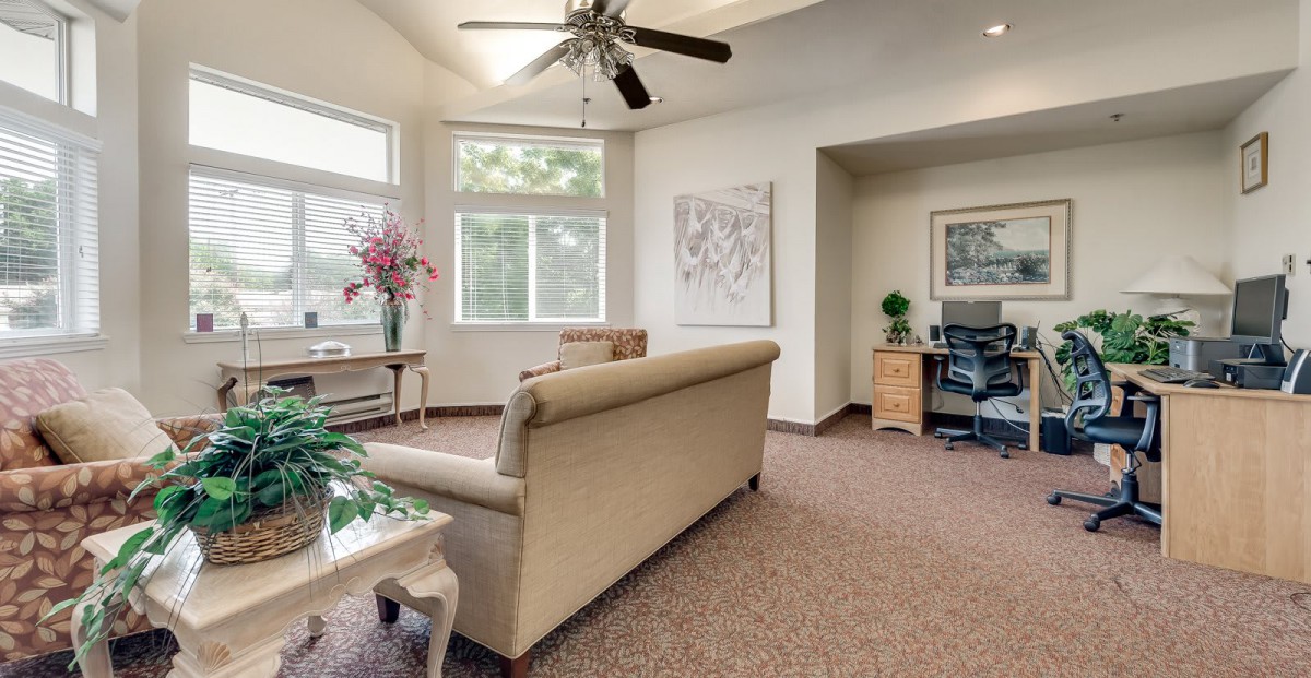 A cozy living room with a sofa, carpet, and ceiling fan, featuring large windows and a small desk area.